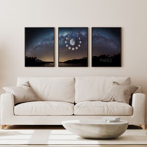 May include: Three framed art prints depicting a night sky with the Milky Way and the phases of the moon. The prints are black framed and hang above a white sofa. The text "MOON" and "PHASES" are printed on the bottom of two of the prints.