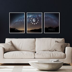May include: Three framed art prints depicting a night sky with the Milky Way. The center print shows the phases of the moon. The left print says "MOON" and the right print says "PHASES". The prints are displayed above a beige sofa and a concrete coffee table.