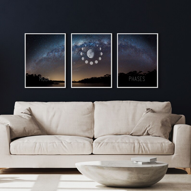 May include: Three framed art prints depicting a night sky with the Milky Way. The center print shows the phases of the moon. The left print says "MOON" and the right print says "PHASES". The prints are displayed above a beige sofa and a concrete coffee table.