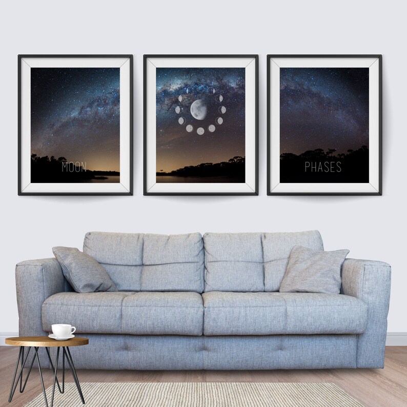 May include: Three framed art prints featuring celestial themes. The prints depict the night sky with the Milky Way, moon phases, and the text "MOON" and "PHASES". The frames are black, and the prints are displayed above a gray sofa.