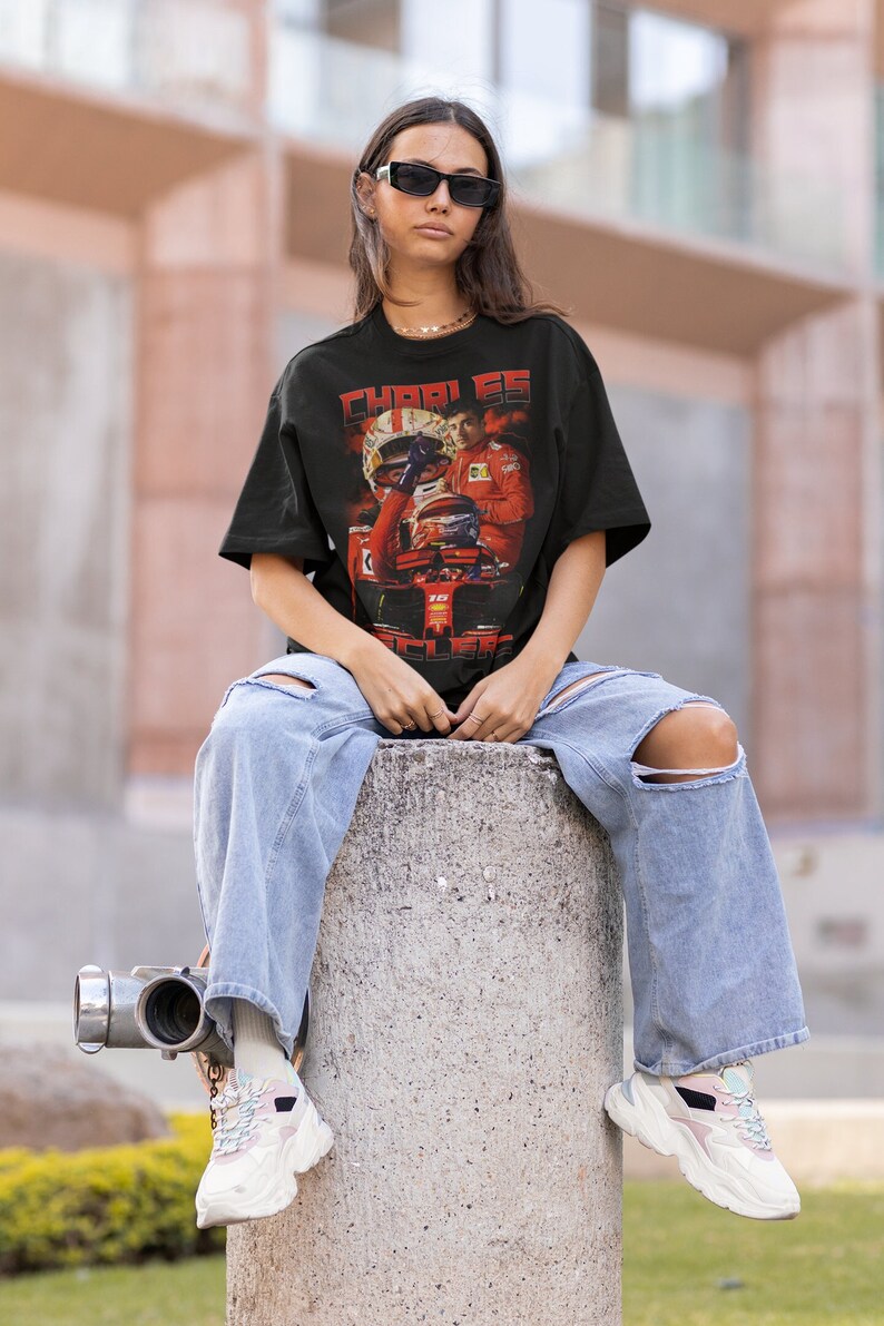 May include: Black graphic t-shirt featuring a vintage-style image of a Formula 1 driver. The shirt has the name "CHARLES LECLERC" in red lettering. The person is wearing light blue ripped jeans and white sneakers.