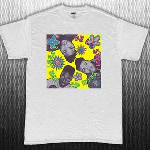 Puede incluir: Camiseta blanca con un diseño gráfico con cuatro caras y el texto "De La Soul" y "Feet High and Rising" sobre un fondo amarillo con ilustraciones de flores. La camiseta está hecha de un material suave.