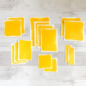 May include: A collection of yellow square and rectangular stickers with white borders, arranged on a light grey and white wooden surface. The stickers are in various sizes and shapes, with a bright, solid yellow colour.