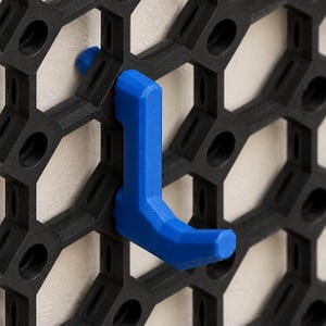 May include: A bright blue L-shaped hook attached to a black hexagonal grid panel. The hook features a faceted, geometric design and is securely mounted into one of the circular openings of the interlocking honeycomb-patterned wall storage system.
