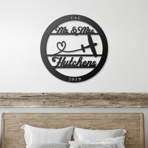 May include: A black metal sign with the text "EST 2019 Mr. & Mrs. Hutchens" and a small airplane with a heart above it. The sign is hanging on a white wall above a wooden headboard.