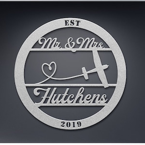 May include: A silver metal circle with the text "EST 2019" at the bottom, "Mr. & Mrs. Hutchens" in a script font, and a small heart and a silhouette of a glider plane in the center.