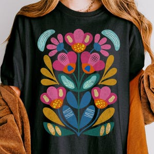 May include: Black t-shirt featuring a vibrant floral design. The graphic showcases pink, blue, and yellow flowers with green leaves and stems. The shirt is a comfortable, casual style, suitable for everyday wear.