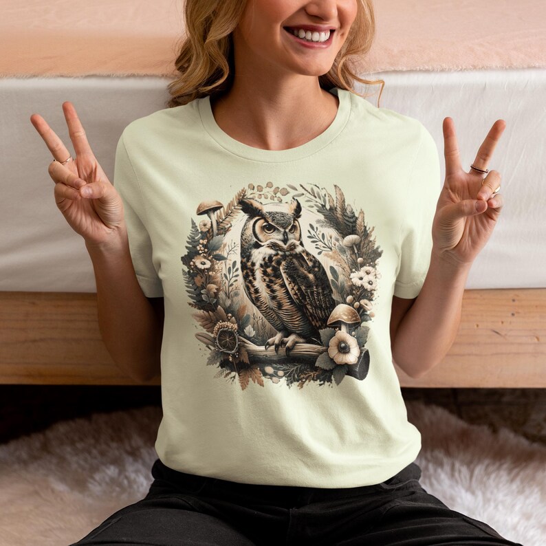 May include: A cream-coloured t-shirt featuring a graphic of an owl perched on a branch surrounded by leaves, mushrooms, and flowers. The owl is brown and black with yellow eyes.