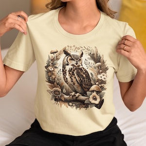 May include: A cream coloured t-shirt with a graphic print of an owl perched on a branch surrounded by mushrooms and flowers.