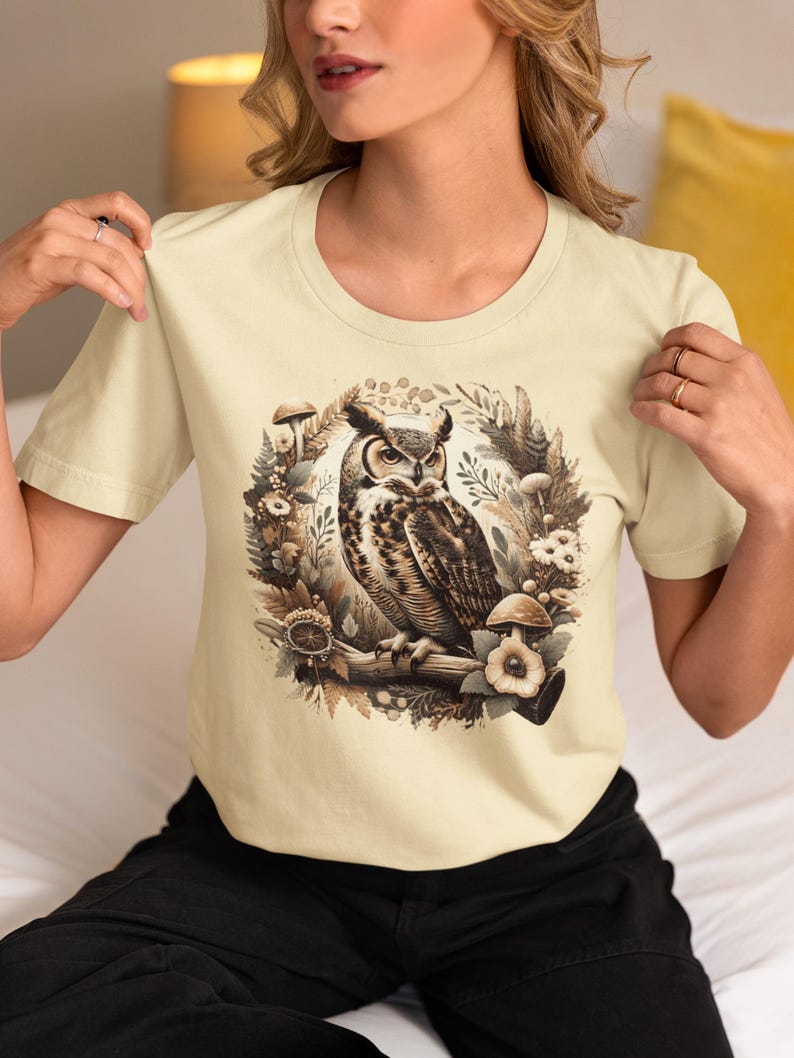May include: A cream coloured t-shirt with a graphic print of an owl perched on a branch surrounded by mushrooms and flowers.