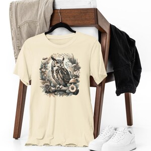 May include: A beige t-shirt featuring a graphic print of an owl perched on a branch surrounded by mushrooms and foliage.