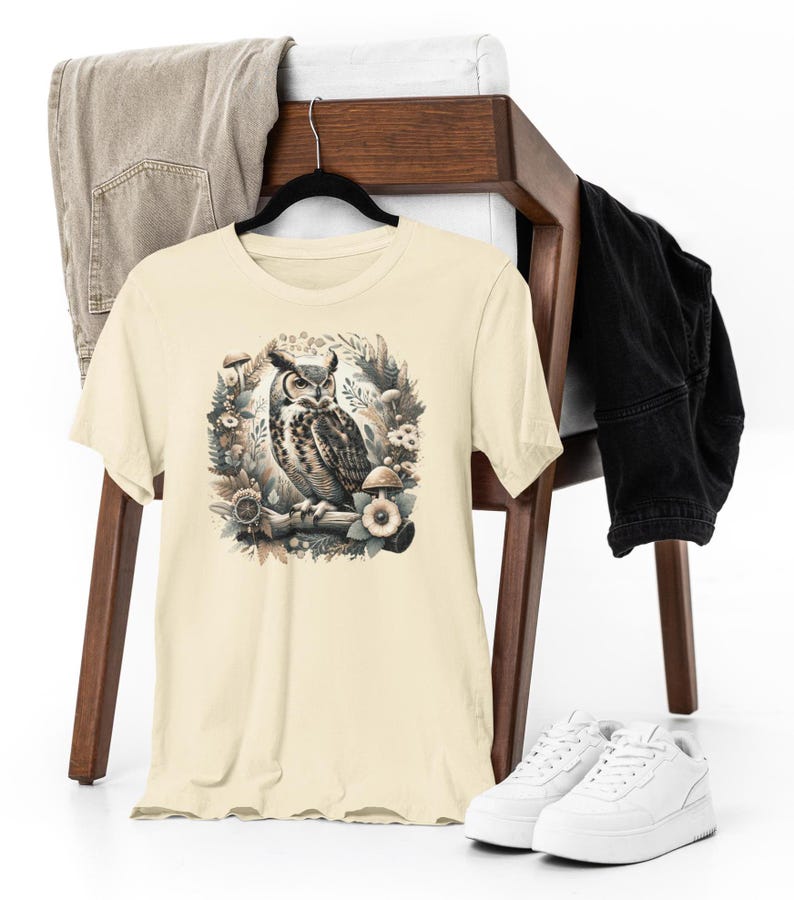 May include: A beige t-shirt featuring a graphic print of an owl perched on a branch surrounded by mushrooms and foliage.