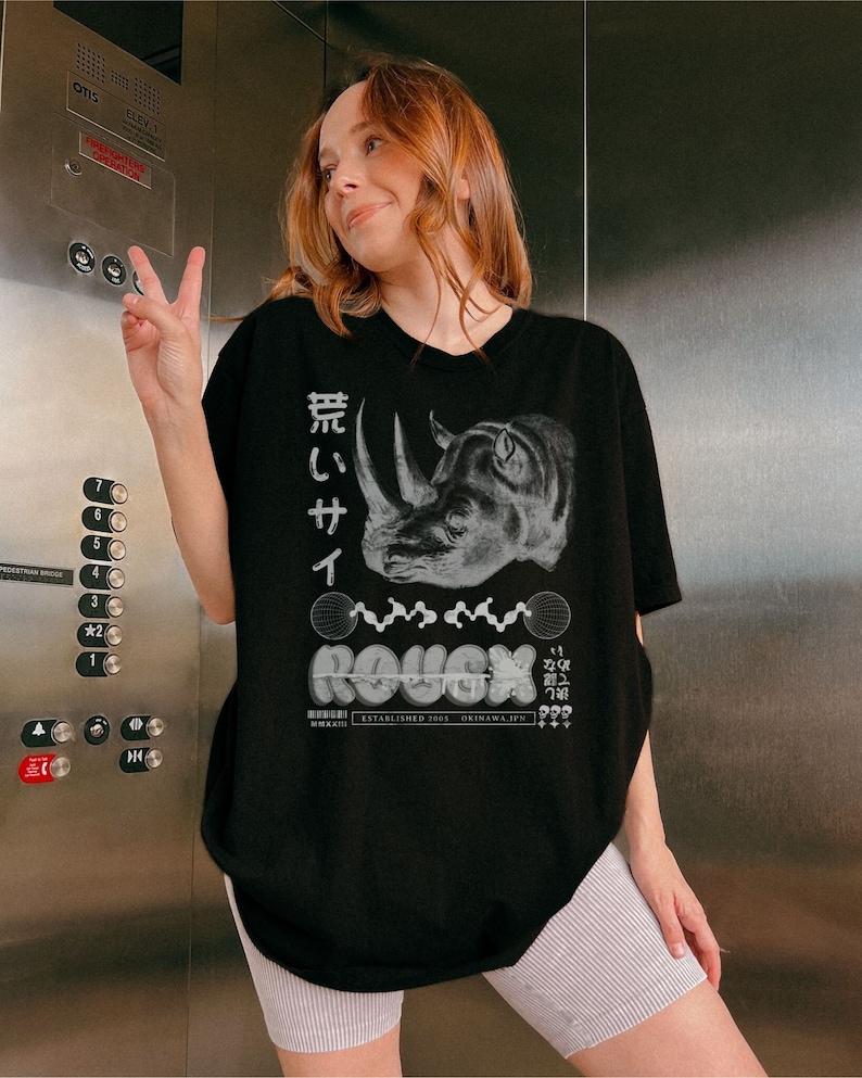 May include: Black oversized t-shirt with a graphic print of a rhinoceros, Japanese text, and the word "ROUGH" in a stylized font. The text "ESTABLISHED 2005 OKINAWA, JPN" is also printed on the shirt.