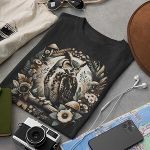 May include: A black t-shirt featuring a graphic print of an owl surrounded by flowers and mushrooms. The owl is brown and white with yellow eyes. The flowers are white and pink. The mushrooms are brown and white.