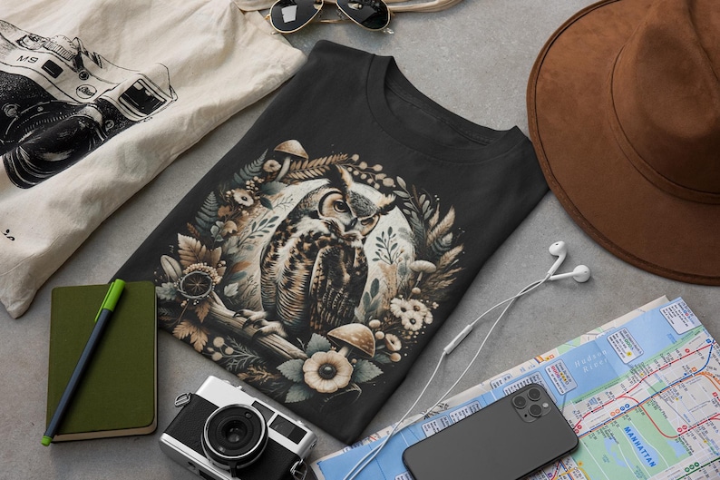 May include: A black t-shirt featuring a graphic print of an owl surrounded by flowers and mushrooms. The owl is brown and white with yellow eyes. The flowers are white and pink. The mushrooms are brown and white.