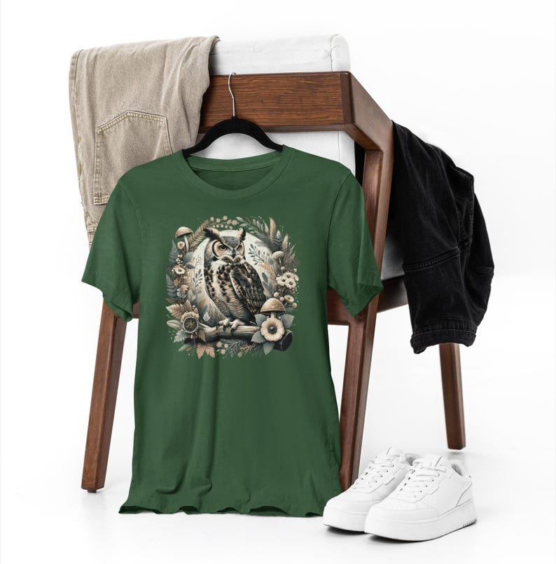 May include: A green t-shirt featuring a graphic of an owl perched in a woodland setting. The owl is brown and white with yellow eyes. The woodland scene includes leaves, branches, and mushrooms.