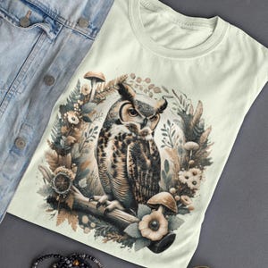 May include: A light green t-shirt featuring a brown and white owl illustration surrounded by mushrooms and flowers. The owl is perched on a branch with its wings spread out.