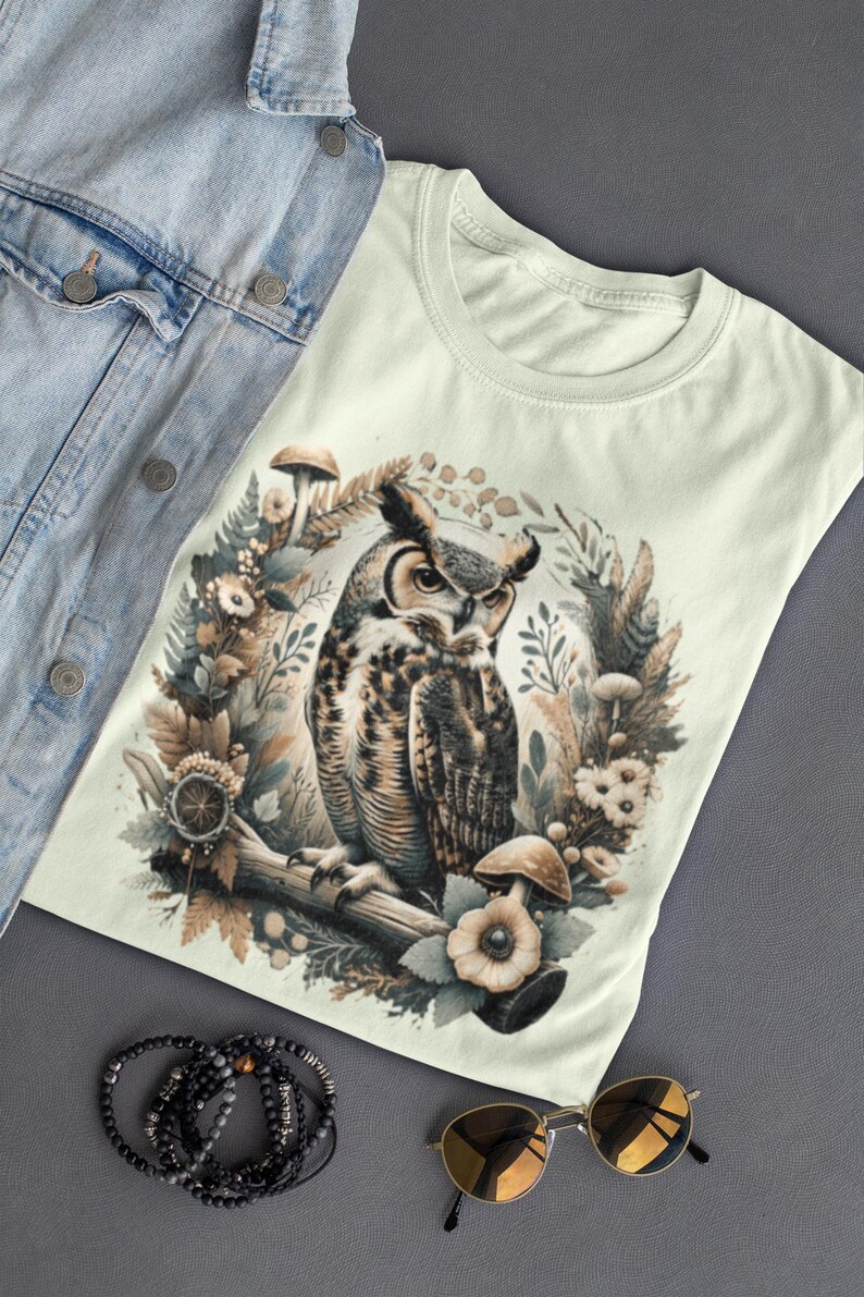 May include: A light green t-shirt featuring a brown and white owl illustration surrounded by mushrooms and flowers. The owl is perched on a branch with its wings spread out.