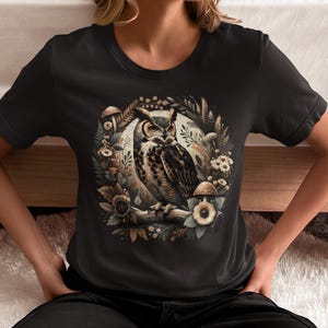 May include: A black t-shirt featuring a graphic print of an owl surrounded by mushrooms and foliage. The owl is brown and white with yellow eyes. The background is black.