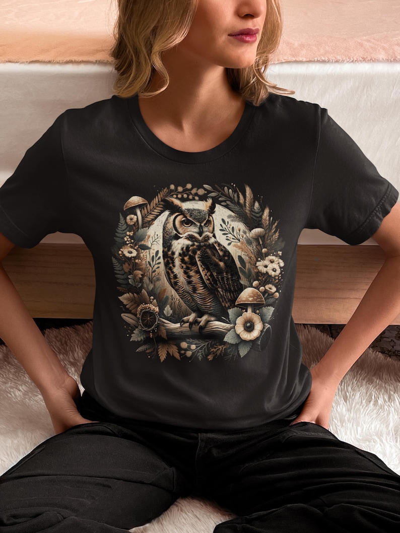 May include: A black t-shirt featuring a graphic print of an owl surrounded by mushrooms and foliage. The owl is brown and white with yellow eyes. The background is black.