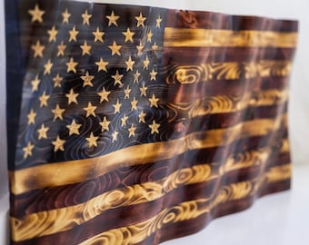 Puede incluir: Una bandera americana de madera con un diseño ondulado. La bandera presenta las estrellas y las rayas tradicionales, con un campo azul oscuro para las estrellas y rayas marrones y beige alternas. Las estrellas son doradas.