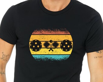 May include: A black t-shirt featuring a retro-style graphic of a pickleball paddle and ball against a sunset backdrop.