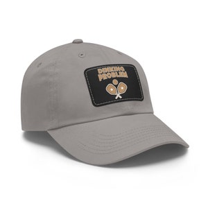 Puede incluir: Una gorra de béisbol gris con un parche negro que dice "DINKING PROBLEM" con un gráfico de dos palas de pickleball y una pelota.