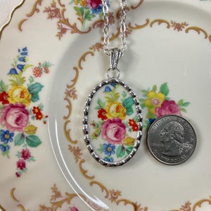 May include: An oval pendant necklace featuring a floral pattern of pink, yellow, and blue flowers on a white background, set in a silver-toned scalloped frame on a silver chain, shown next to a U.S. quarter for scale to indicate its size.