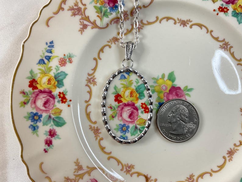May include: An oval pendant necklace featuring a floral pattern of pink, yellow, and blue flowers on a white background, set in a silver-toned scalloped frame on a silver chain, shown next to a U.S. quarter for scale to indicate its size.