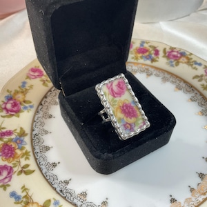 May include: A rectangular ring featuring a floral pattern with pink, yellow, and blue flowers on a white background, set in a silver-toned scalloped metal frame. The ring is displayed inside a black velvet jewelry box with a hinged lid.