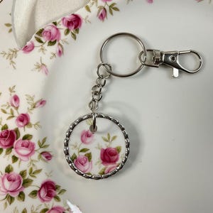 May include: A circular keychain featuring a white ceramic insert decorated with pink roses and green leaves, encased in a scalloped silver-tone metal frame. The keychain includes a silver-tone metal chain and a lobster claw clasp attachment.