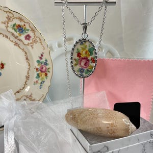 May include: A silver-toned necklace featuring an oval pendant with a floral pattern of pink, yellow, and blue flowers. The pendant hangs from a delicate silver chain. Included are a beige seashell, a pink cloth, and a white gift box.