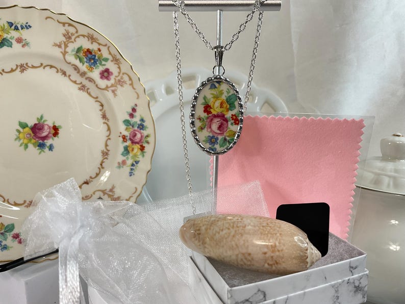 May include: A silver-toned necklace featuring an oval pendant with a floral pattern of pink, yellow, and blue flowers. The pendant hangs from a delicate silver chain. Included are a beige seashell, a pink cloth, and a white gift box.