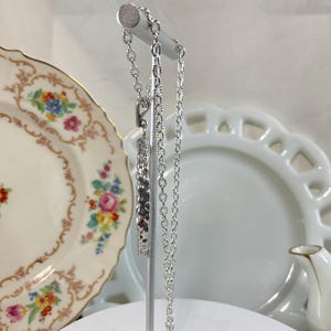 May include: A silver-toned metal chain necklace featuring a rectangular pendant with a textured, repeating geometric pattern. The necklace is displayed on a vertical stand, showcasing the link chain design and the metallic finish of the pendant.
