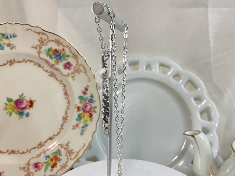 May include: A silver-toned metal chain necklace featuring a rectangular pendant with a textured, repeating geometric pattern. The necklace is displayed on a vertical stand, showcasing the link chain design and the metallic finish of the pendant.