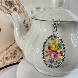 May include: An oval pendant necklace featuring a floral pattern with pink, yellow, and blue flowers on a cream background, encased in a scalloped silver-toned metal frame, suspended from a silver-toned chain against a white teapot.
