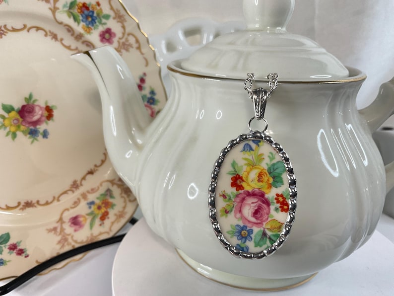 May include: An oval pendant necklace featuring a floral pattern with pink, yellow, and blue flowers on a cream background, encased in a scalloped silver-toned metal frame, suspended from a silver-toned chain against a white teapot.