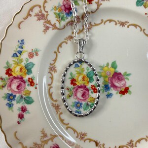 May include: An oval pendant necklace featuring a floral pattern with pink, yellow, and blue flowers on a white background, encased in a scalloped silver-tone metal frame and suspended from a matching silver-tone twisted rope chain link necklace.