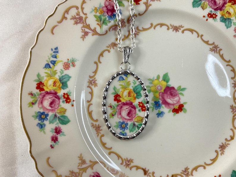 May include: An oval pendant necklace featuring a floral pattern with pink, yellow, and blue flowers on a white background, encased in a scalloped silver-tone metal frame and suspended from a matching silver-tone twisted rope chain link necklace.