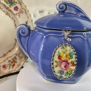 May include: An oval pendant featuring a colorful floral pattern with pink, yellow, and blue flowers on a cream background, encased in a scalloped silver-toned metal frame and suspended from a silver-toned chain against a blue ceramic sugar bowl.