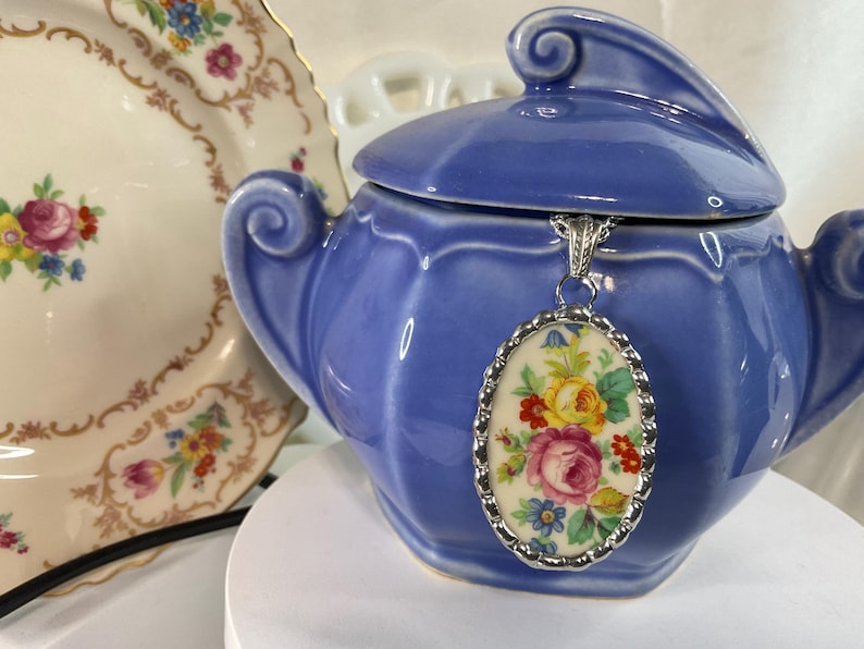 May include: An oval pendant featuring a colorful floral pattern with pink, yellow, and blue flowers on a cream background, encased in a scalloped silver-toned metal frame and suspended from a silver-toned chain against a blue ceramic sugar bowl.