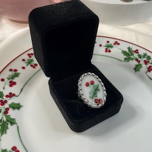 May include: An oval-shaped ring featuring a white center with a painted green holly leaf and red berries, set in a scalloped silver-toned metal frame. The ring is displayed inside a black velvet-lined jewelry box.