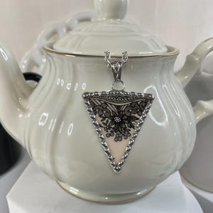 May include: A silver-toned necklace with a triangular pendant featuring a black floral design on a cream background. The pendant is framed with a silver beaded edge. The necklace is displayed on a white teapot.