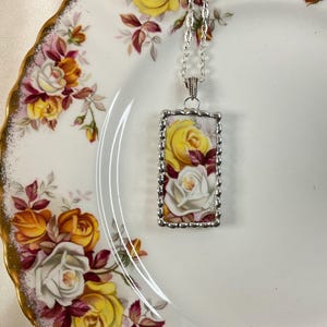 May include: A rectangular pendant necklace featuring a floral pattern of yellow and white roses on a white background, encased in a silver-toned scalloped metal frame and suspended from a matching silver-toned link chain necklace.