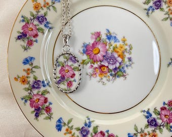May include: A silver necklace with a floral oval pendant. The pendant features a cluster of pink, purple, and yellow flowers on a white background. The necklace is displayed on a white plate with a floral pattern.