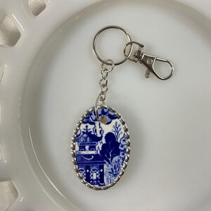 May include: An oval-shaped keychain featuring a blue and white willow pattern design. The pendant is encased in a silver-toned scalloped metal frame and is attached to a silver-toned chain, a circular split ring, and a lobster claw clasp fastener.