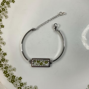 May include: A silver-toned metal bracelet featuring a rectangular center charm with a white and olive green floral pattern. The bracelet includes a smooth curved band, a delicate chain extension, and a small lobster claw clasp closure.