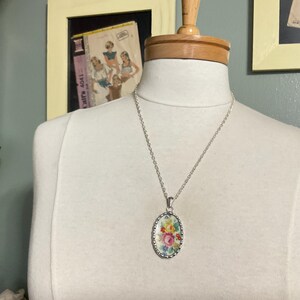 May include: A silver-toned necklace featuring an oval pendant with a scalloped edge. The pendant displays a colorful floral pattern with pink, yellow, and green flowers on a white background, suspended from a simple silver-toned metal chain.