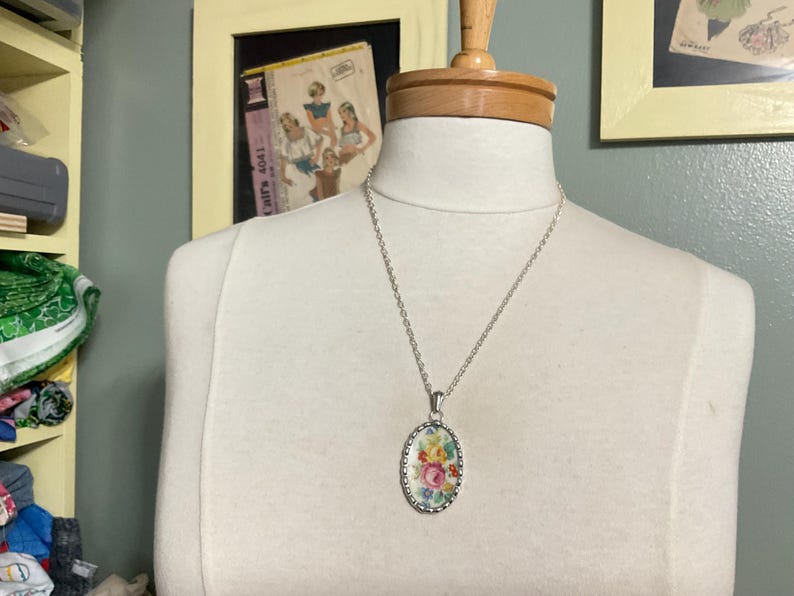 May include: A silver-toned necklace featuring an oval pendant with a scalloped edge. The pendant displays a colorful floral pattern with pink, yellow, and green flowers on a white background, suspended from a simple silver-toned metal chain.
