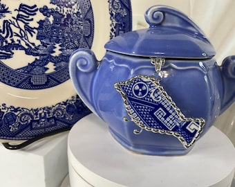 May include: A blue ceramic sugar bowl with a lid, featuring a decorative silver-toned pendant. The pendant has a blue and white design. A blue and white plate is in the background.